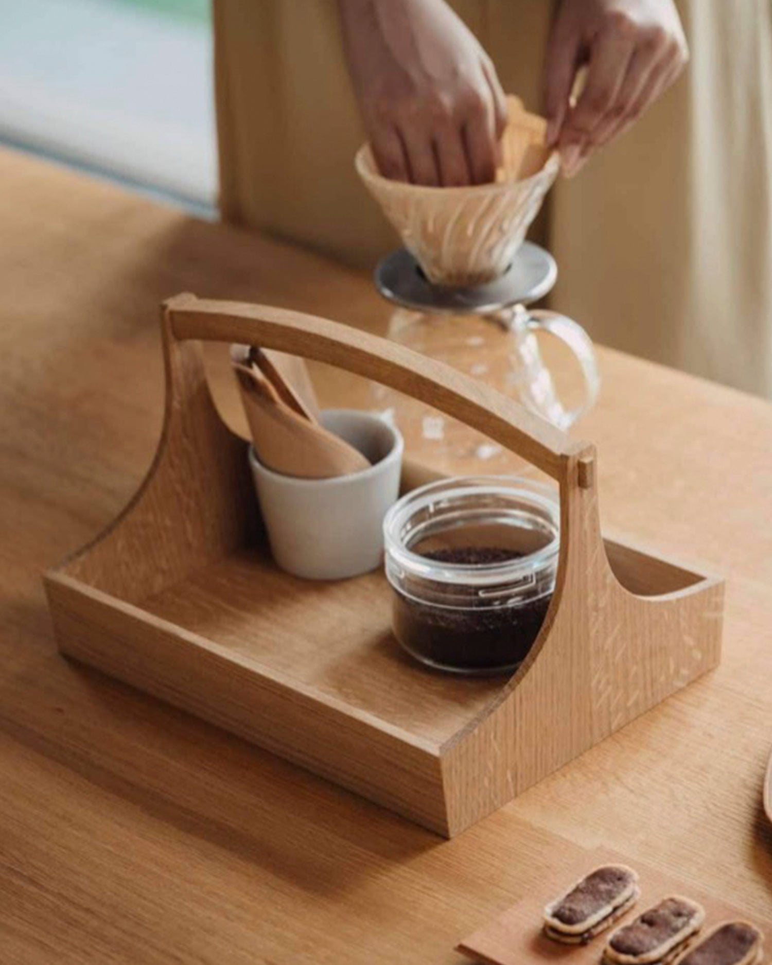 Person making coffee using a pour-over method on large wooden table caddy by Aizawa with short handles, carrying a jar of coffee beans and a cup with coffee filters