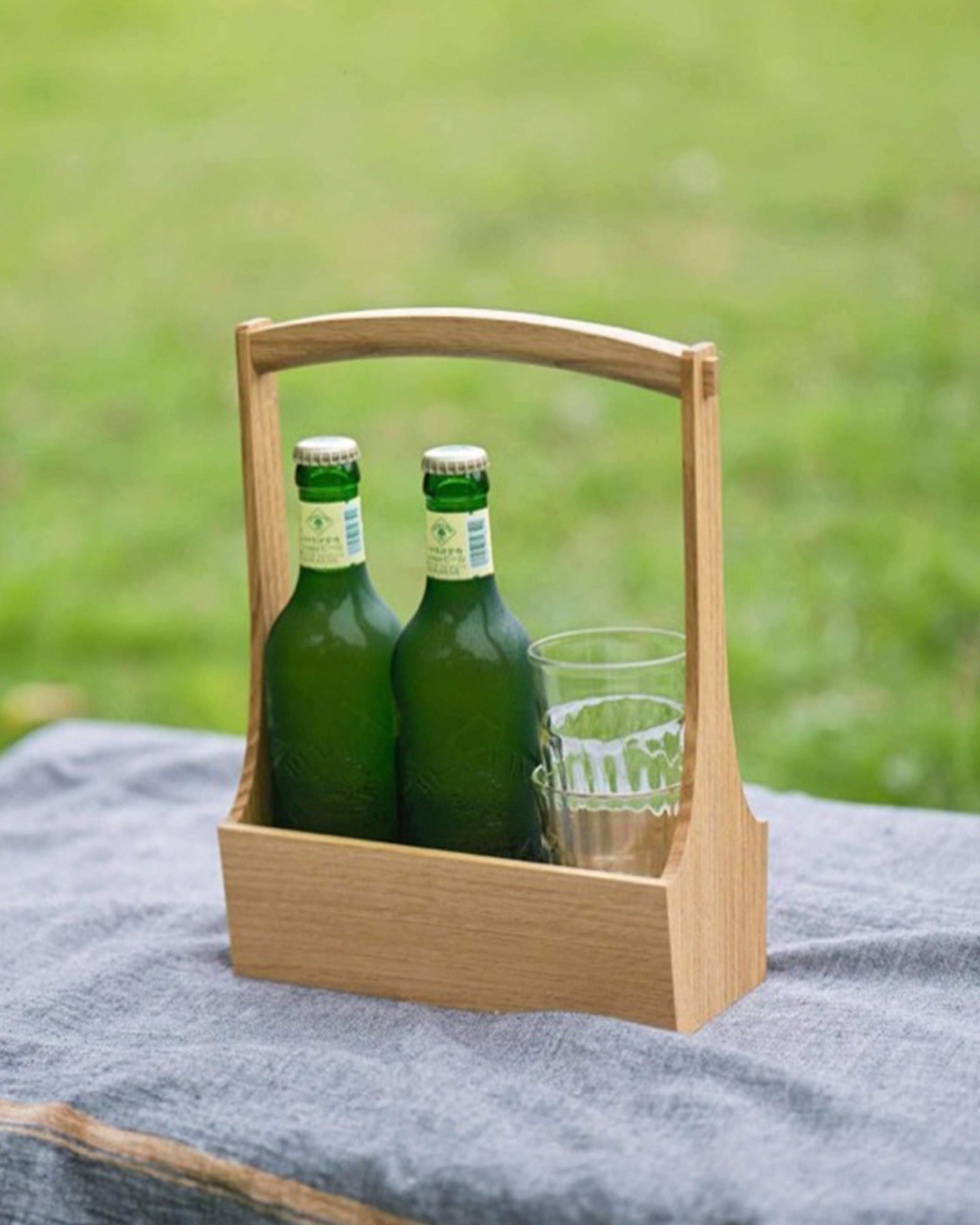 Wooden table caddy by Aizawa with two green bottles and two glass cups stacked on blanket with a grass background.