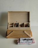 In situation image of open chestnut wood picnic box by Shinsuke Tanabe with the two inner compartments filled with sewing materials and folded fabric sitting in front.