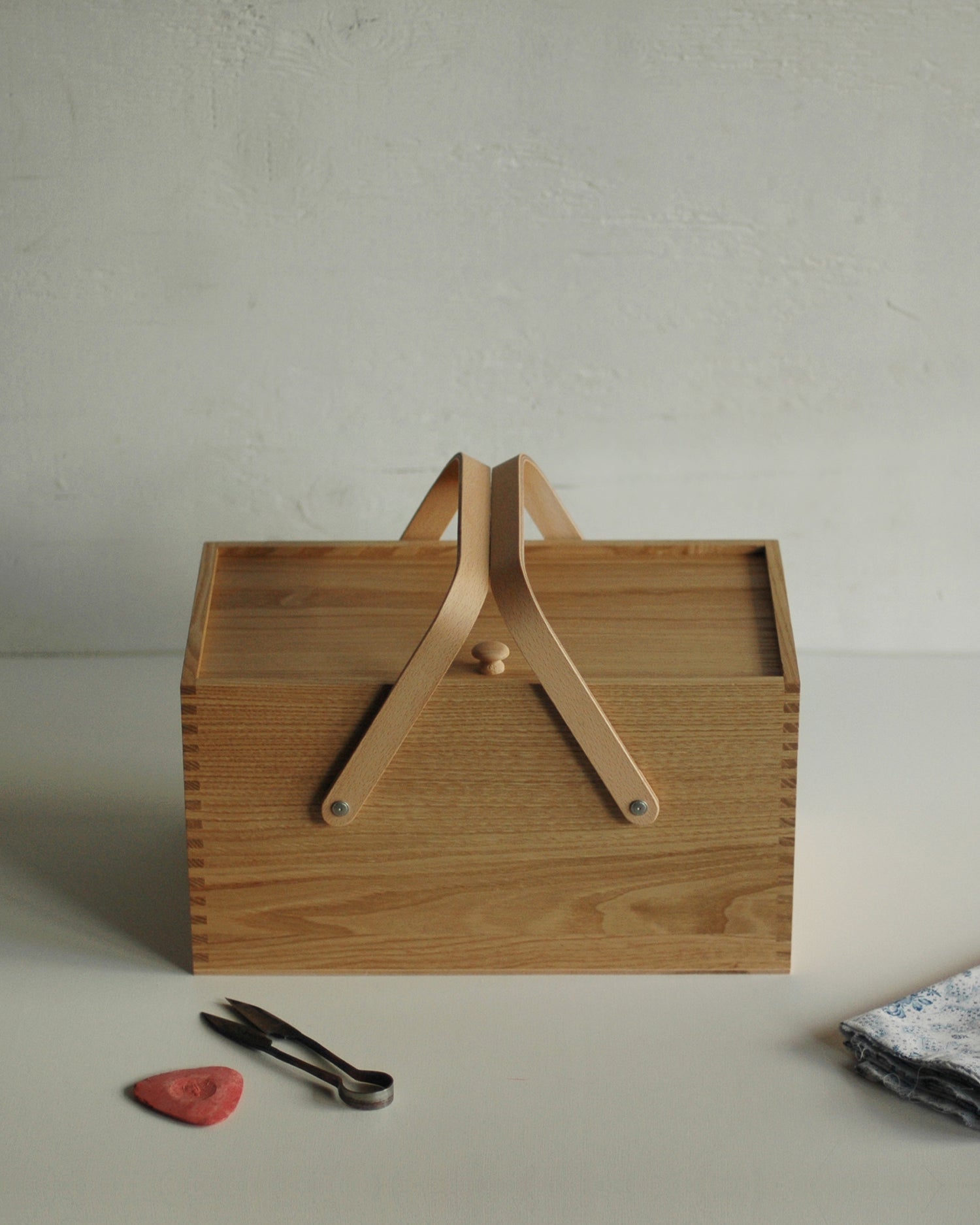 In situation image of chestnut wood picnic box featuring wood joinery on edges by Shinsuke Tanabe next to metal tongs and white napkin with white wall in background.