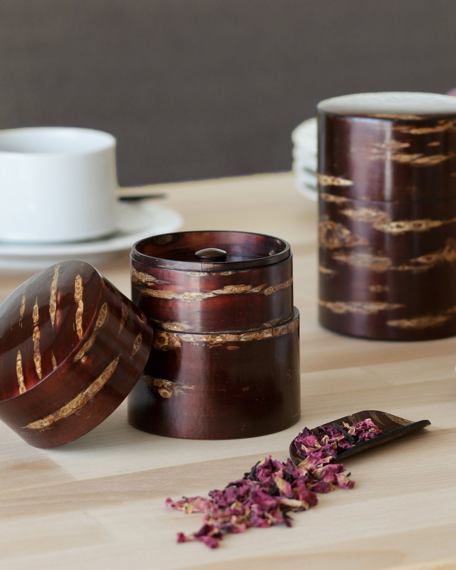 In situation image of Smooth Cherry Bark Scoop by Denshiro with dried rose petals in front of open short and closed tall tea canister on light oak surface with gray wall and white tea cups in the background.