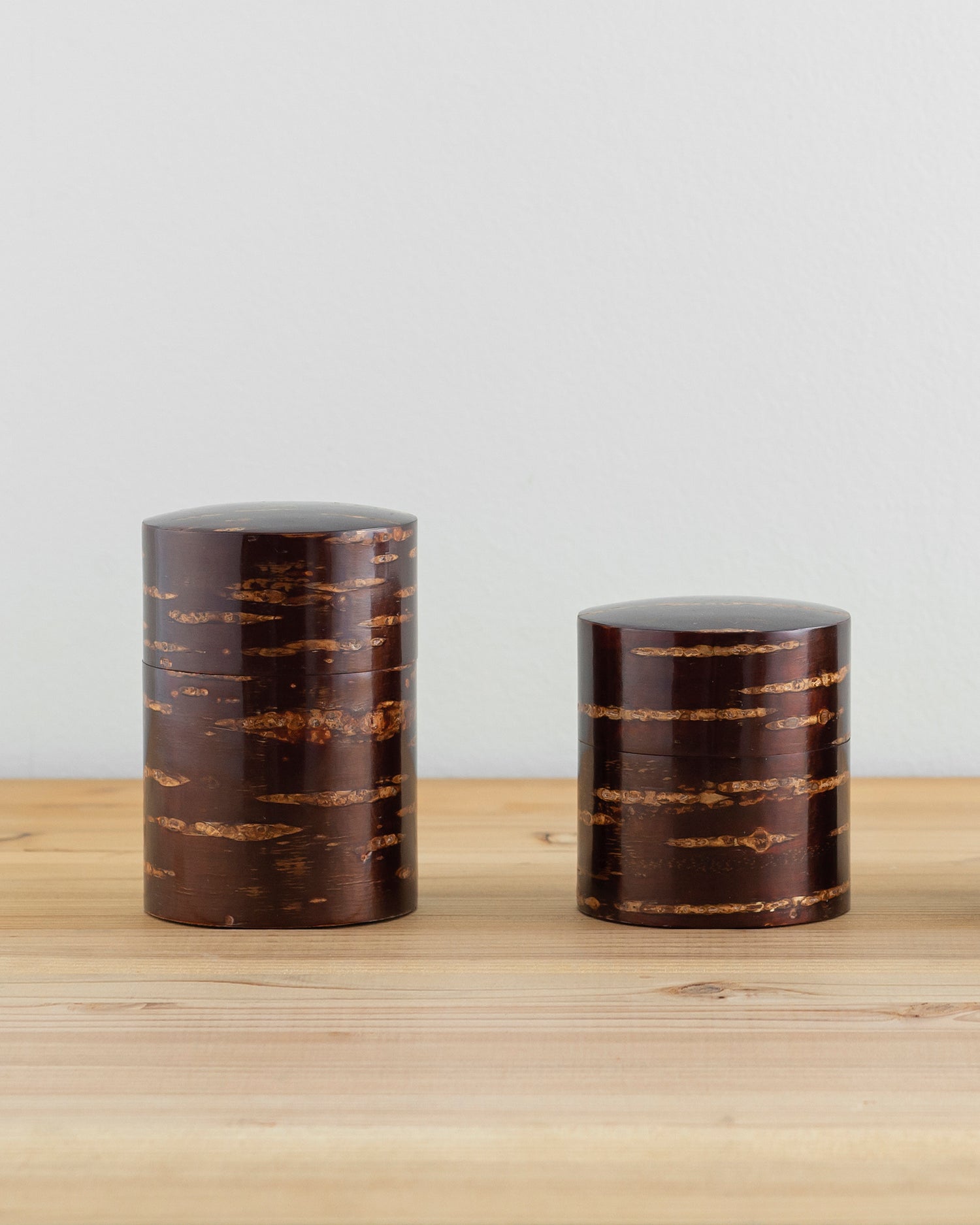 Front view of tall and short Smooth Cherry Bark Tea Canister next to each other on light oak surface with white wall behind.