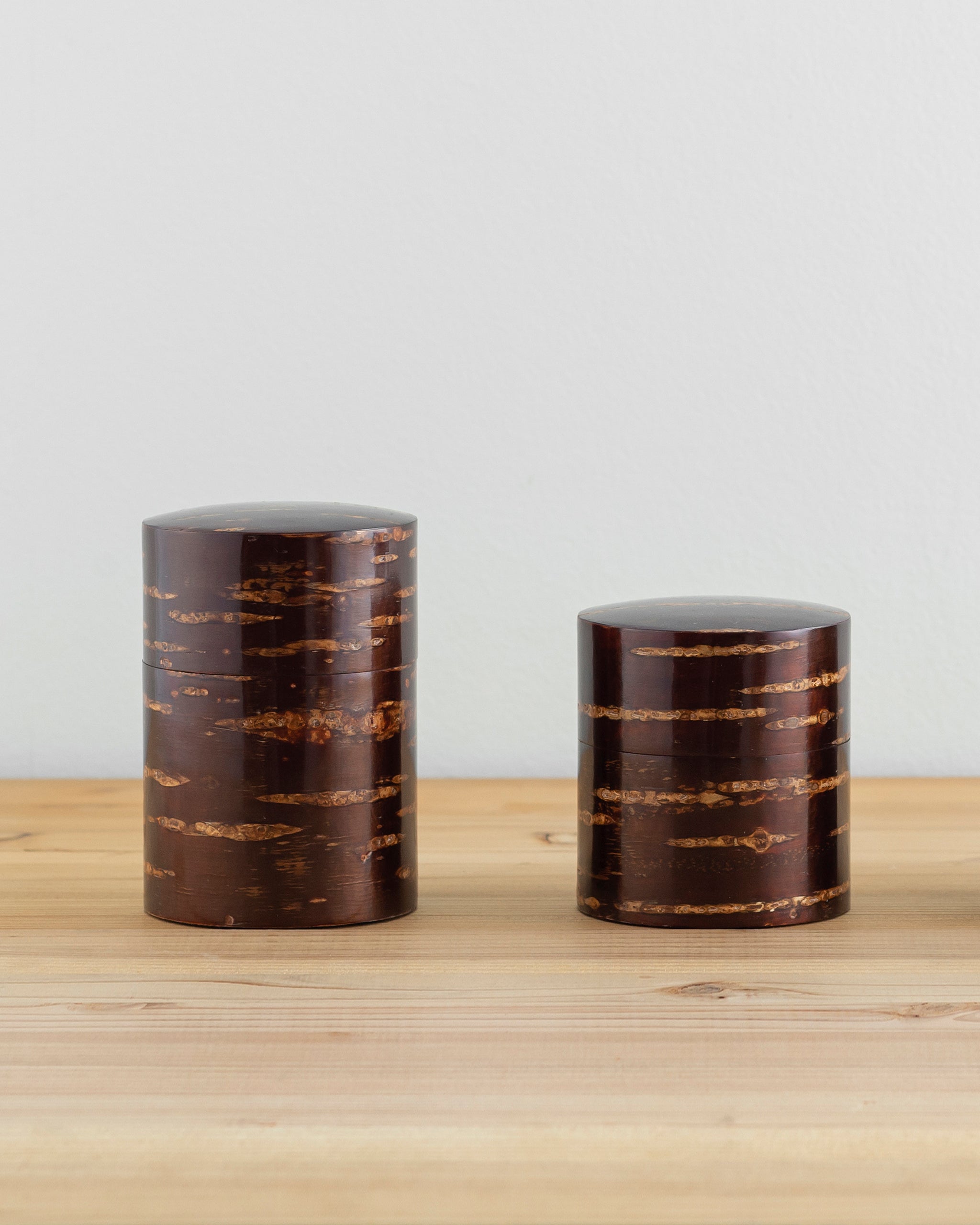 Front view of tall and short Smooth Cherry Bark Tea Canister next to each other on light oak surface with white wall behind.