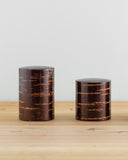 Front view of tall and short Smooth Cherry Bark Tea Canister next to each other on light oak surface with white wall behind.