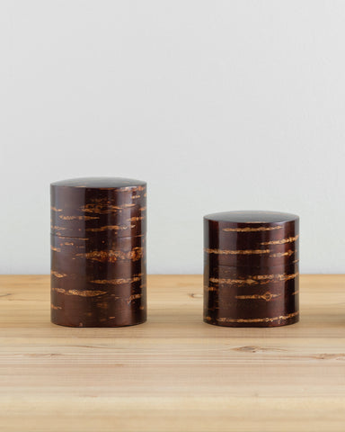 Front view of tall and short Smooth Cherry Bark Tea Canister next to each other on light oak surface with white wall behind.
