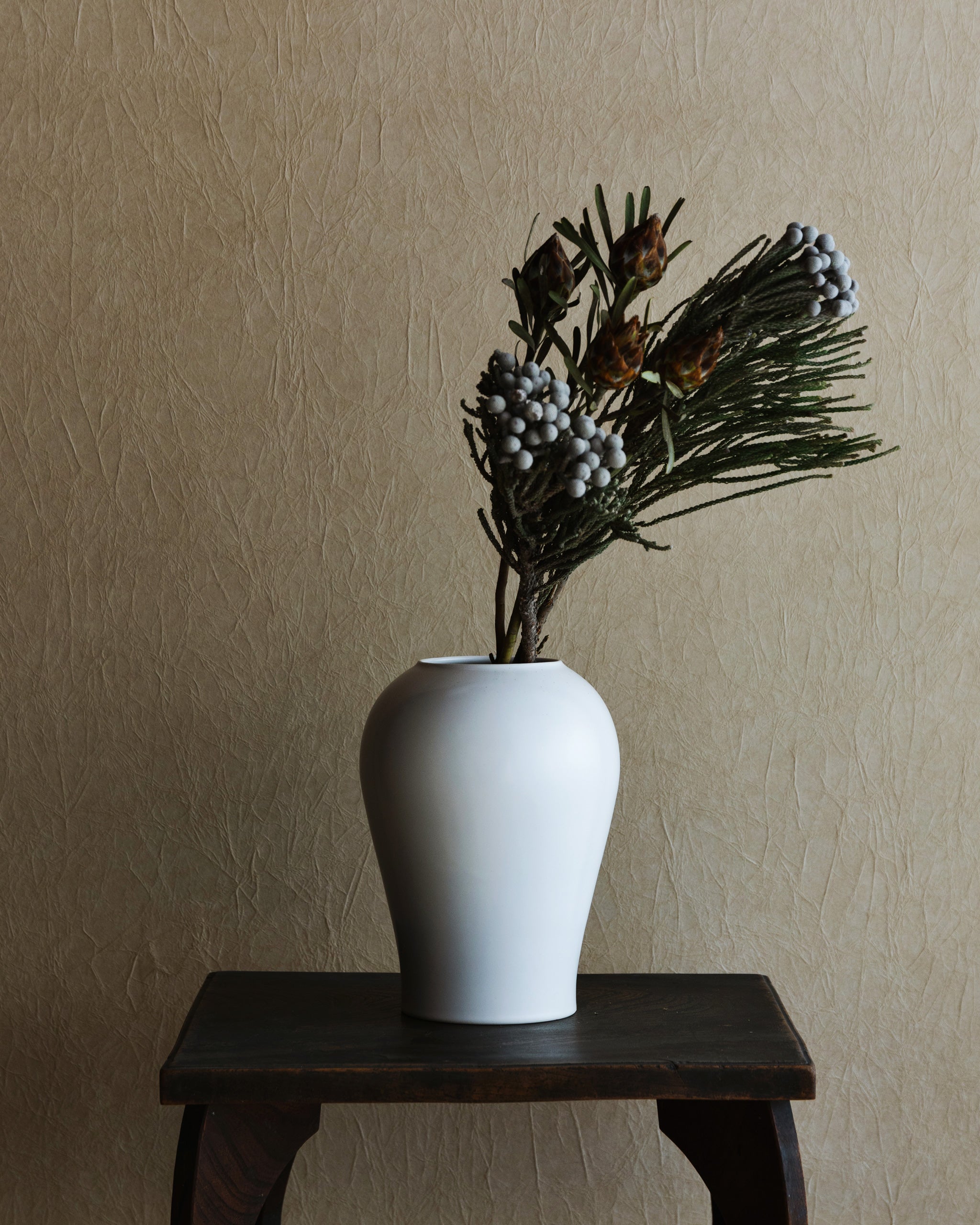 Large heishi vase placed on top of a dark brown wood stool with green branches in it.