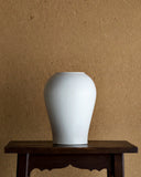 Large heishi vase is on top of a dark brown lacquered stool infront of a textured light brown wall.