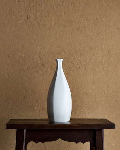 Rokkaku-bin Vase placed on top of a dark brown lacquered stool, against textured light brown wall.