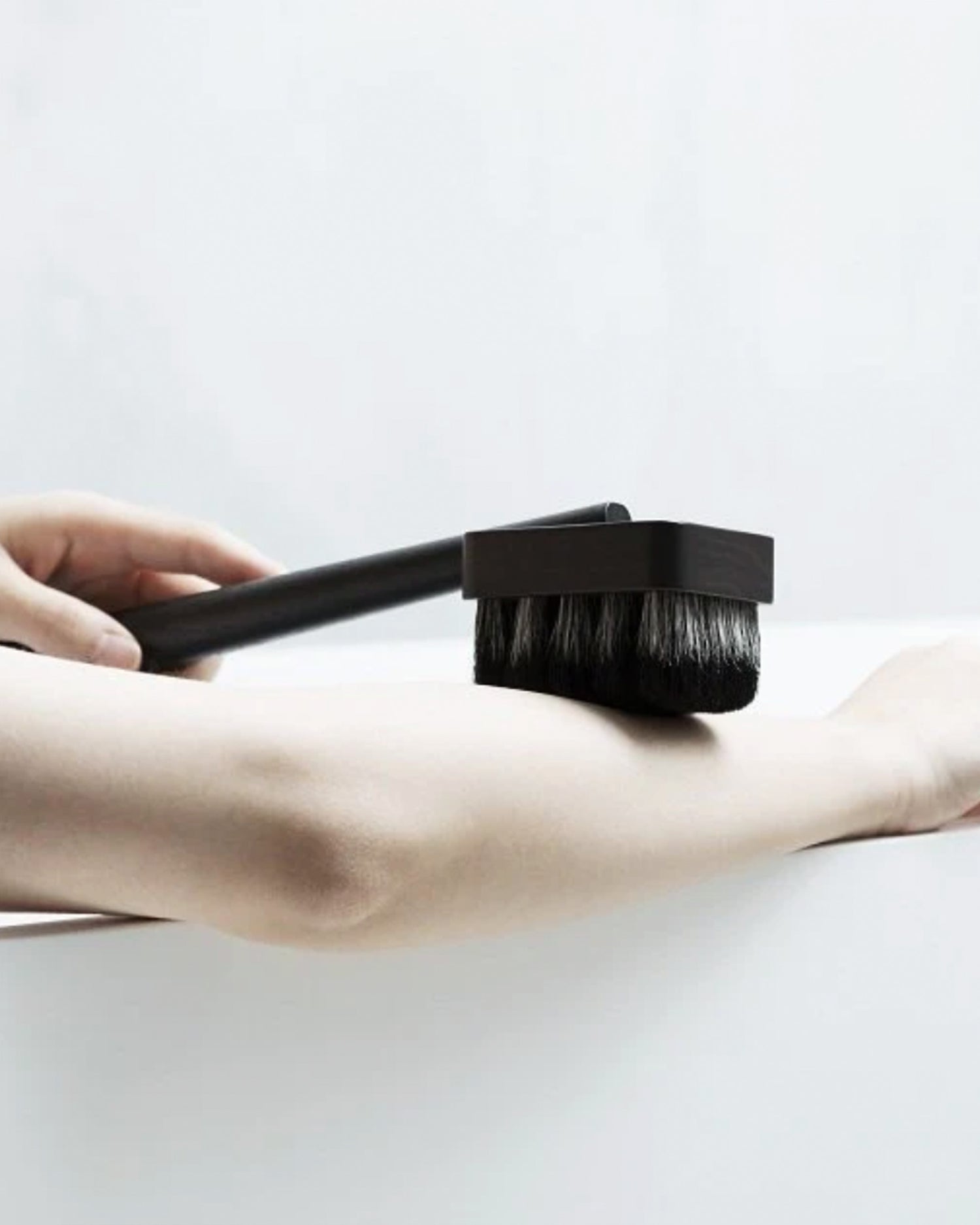 In situation image of hand holding dark long wooden jiva body brush by Shaquda on arm against white background.
