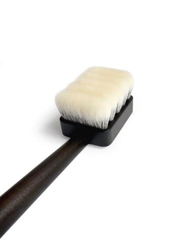 Angled and cropped image of long jiva body brush with dark wood handle and light bristles by Shaquda laying against white background.