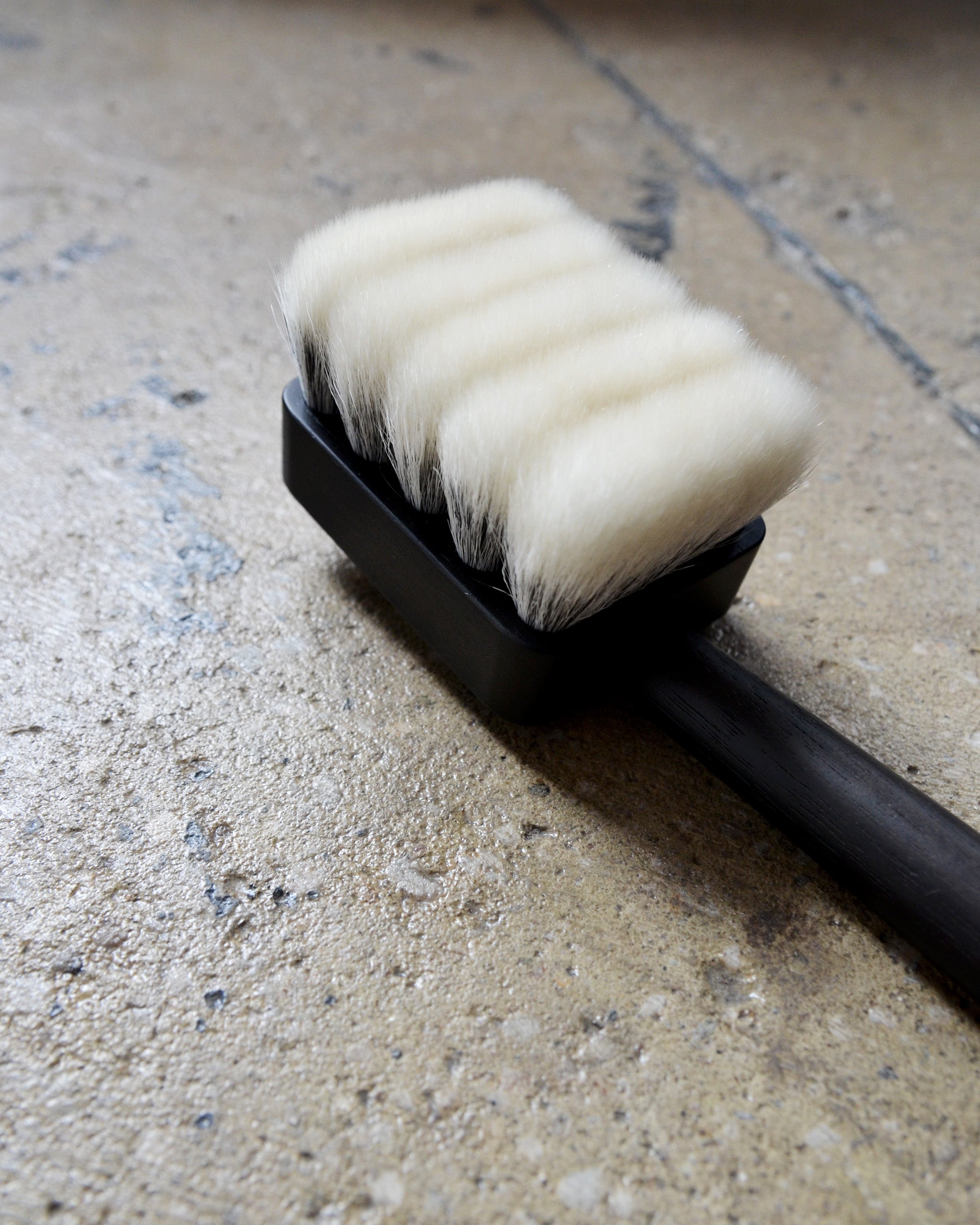 Close-up and cropped image of long jiva body brush with dark wood handle and light bristles by Shaquda against rough cement surface.