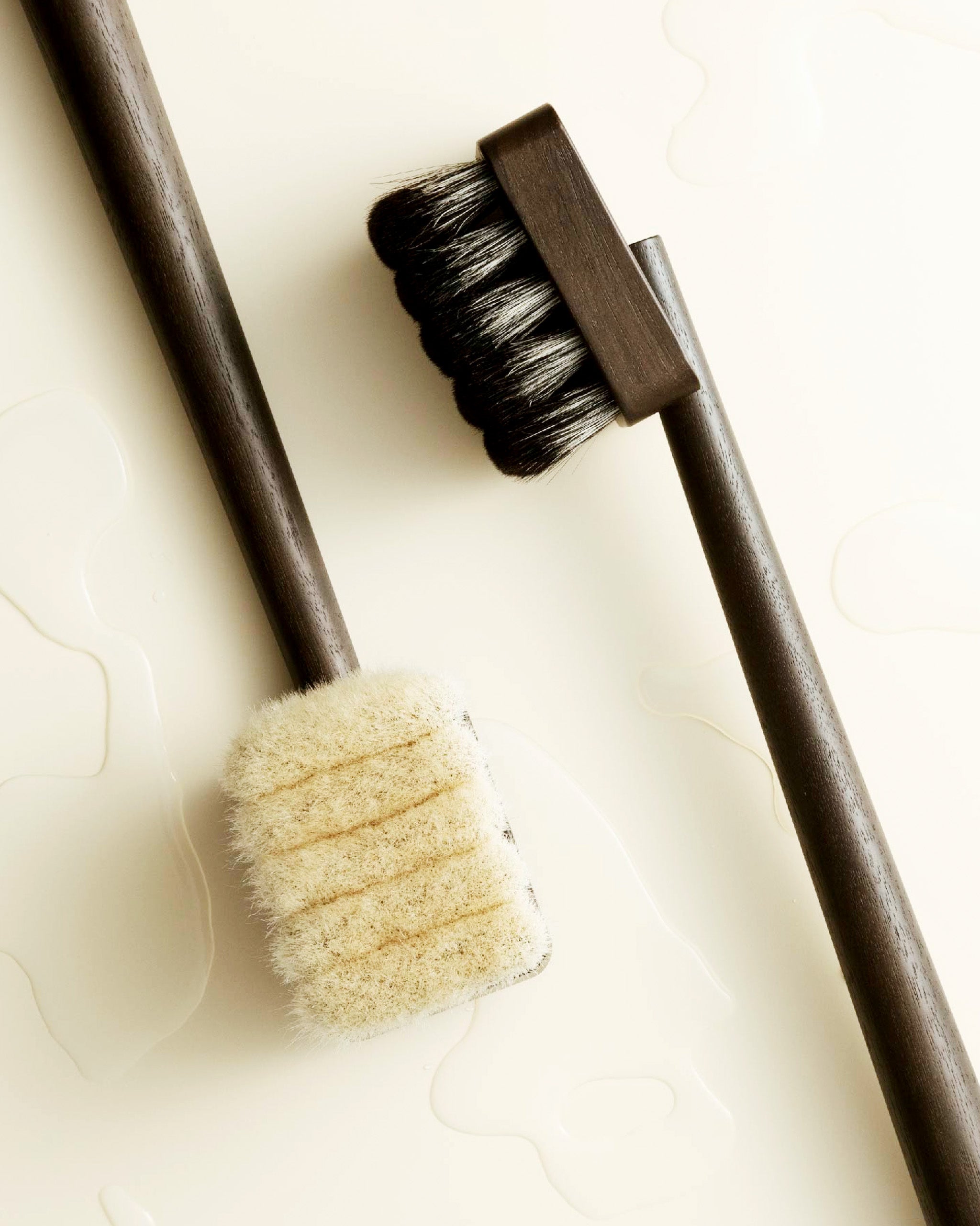 In situation image of dark long wooden jiva body brush by shaquda laying next to light wooden long body brush with water.