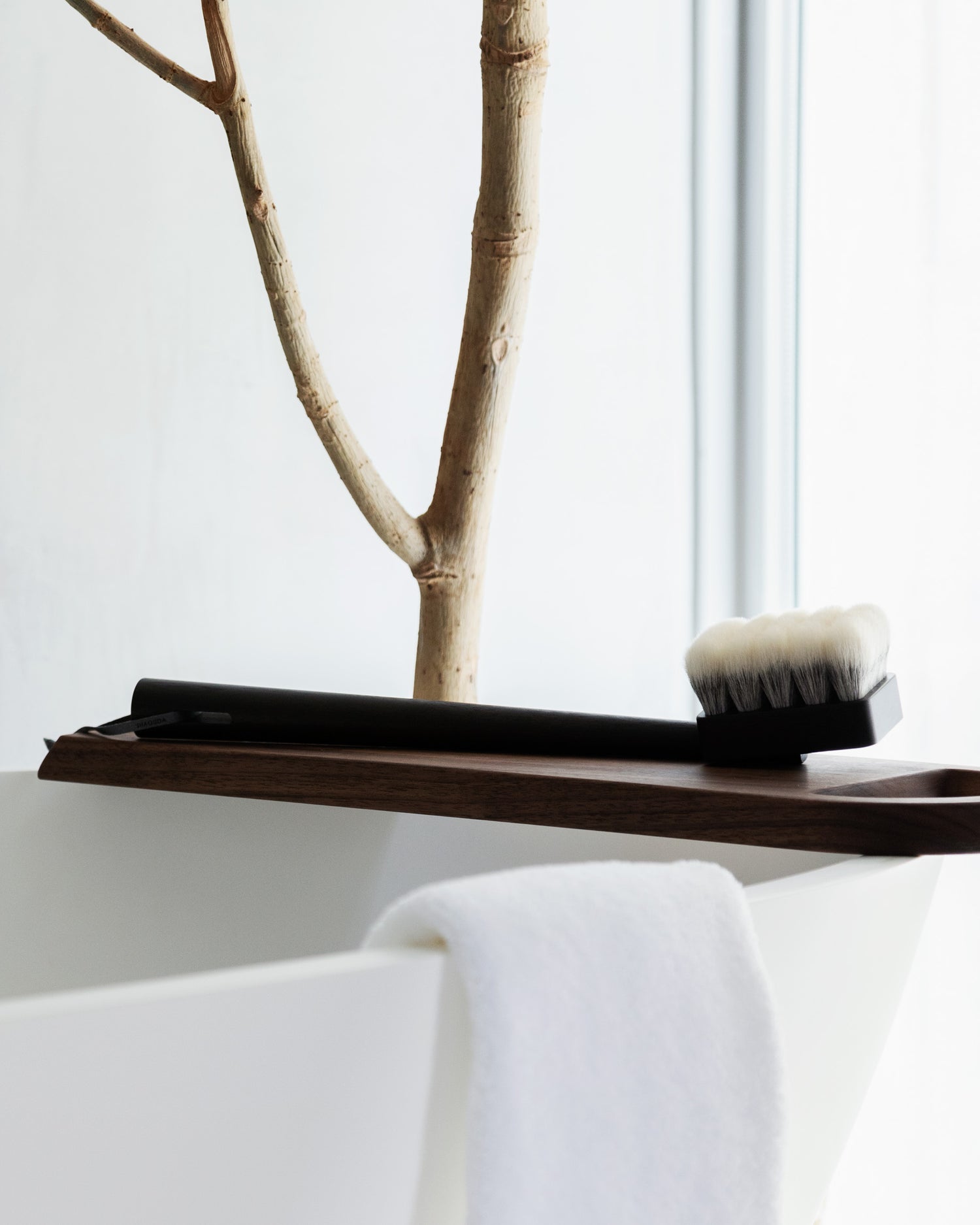 In situation image of long jiva body brush with dark wood handle and dark bristles by Shaquda on wooden bath tray on top of white tub with pale tree in the background.
