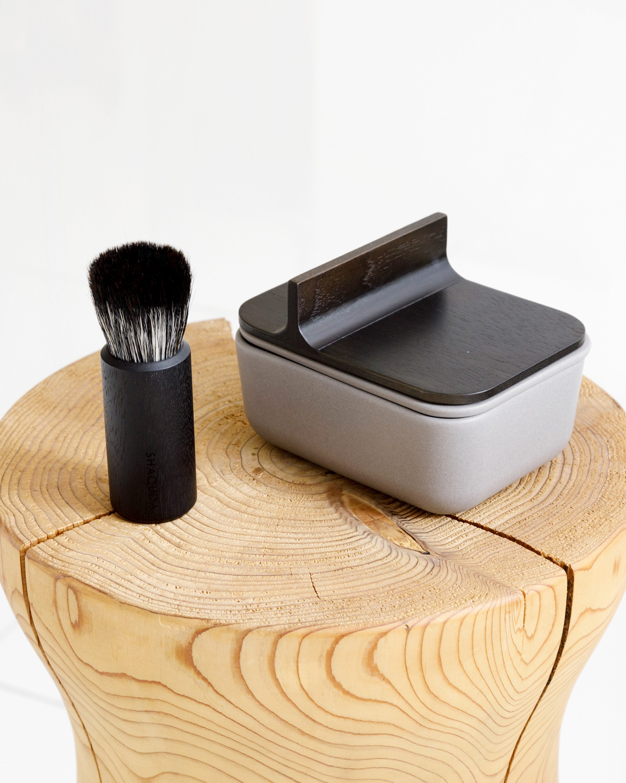 In situation image of hard Jiva face cleansing and shaving series with porcelain bowl and walnut, boar bristle, and goat hair brush by Shaquda on wood stump.