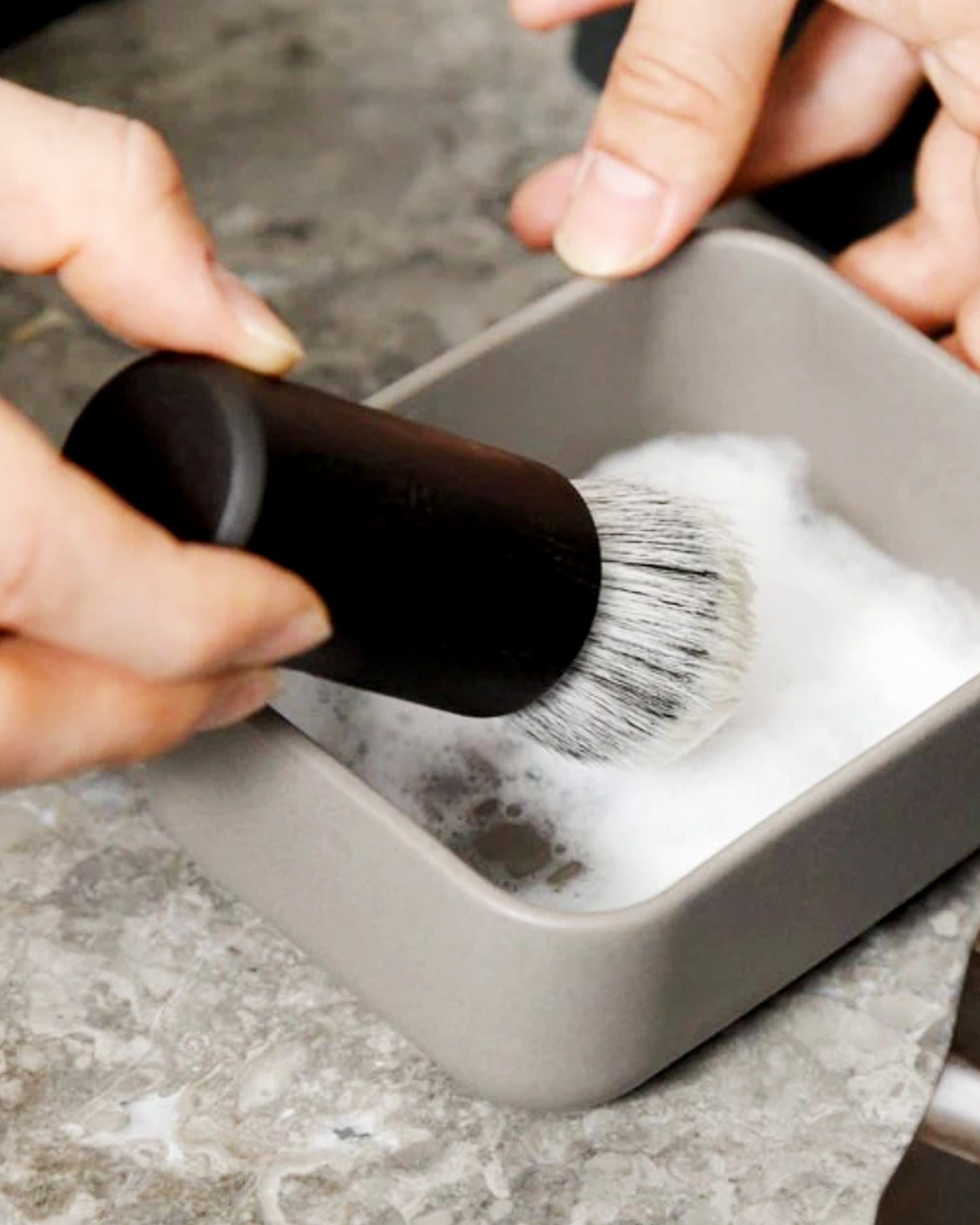 In situation  image of hands using the Jiva Face Cleansing and Shaving series soft brush to mix shaving cream in the porcelain bowl by Shaquda on top of a stone counter. 