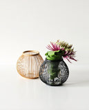 Two round flower baskets. Left one is a natural color while the right one is black. Black basket contains a large folded leaf and a flower in a glass container. The flower is magenta and green.