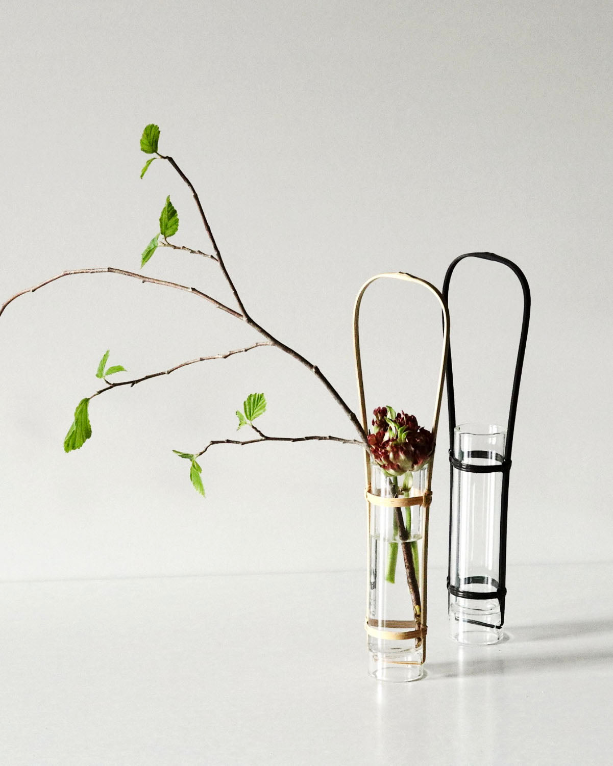 Two slender vases against gray background. Both have bamboo holders that feature a handles so that the user  can hang the vase. The one on the left is natural colored while one on right is black. The natural colored vase has a flower and small tree branch in it.