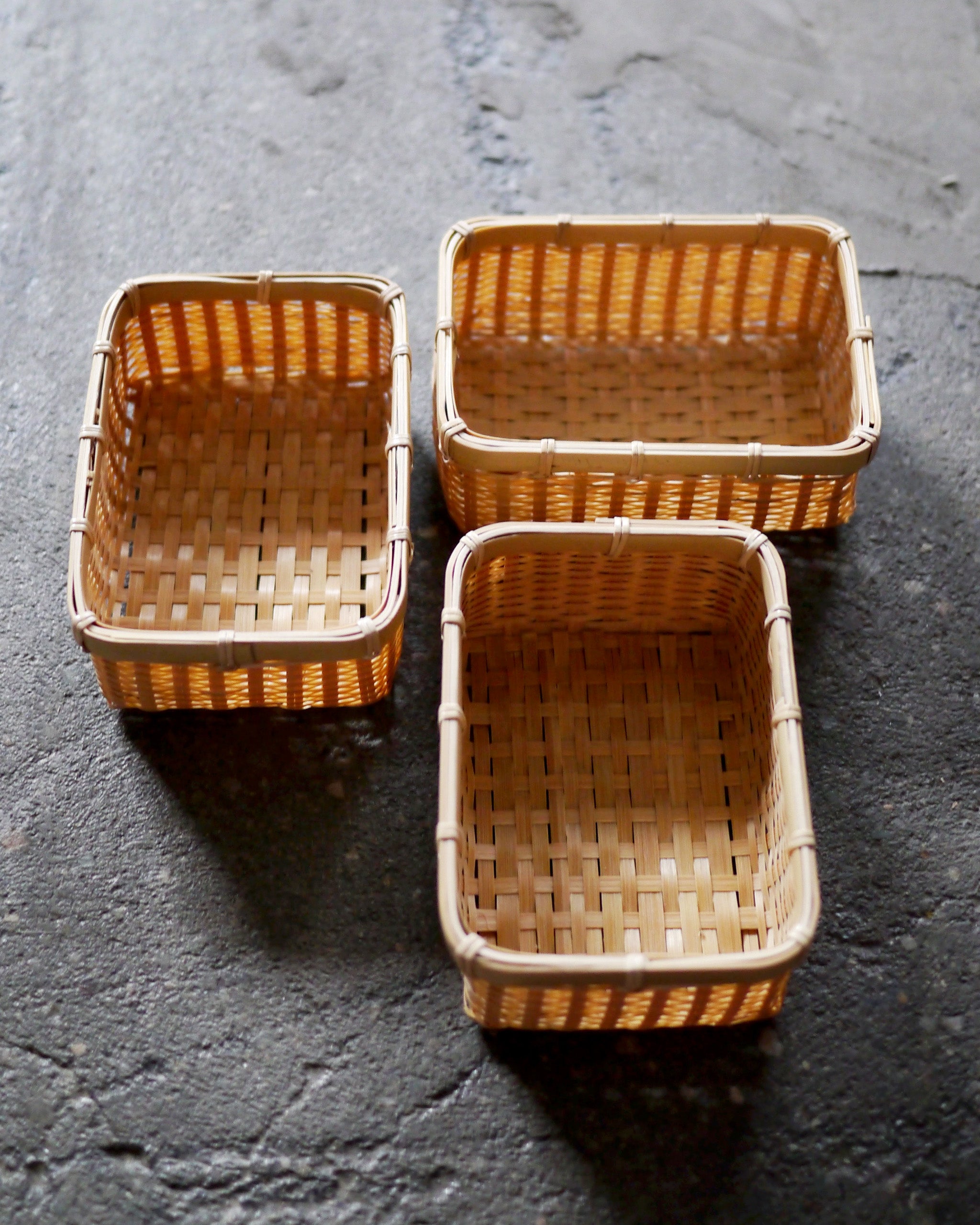Angled in situation image of three small woven rectangular baskets by Kochosai Kosuga on dark cement surface.