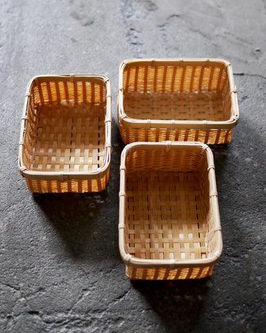 Angled in situation image of three small woven rectangular baskets by Kochosai Kosuga on dark cement surface.
