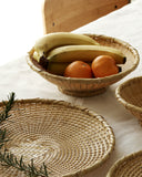 Bowl made out of natural colored woven bamboo sitting against on a beige cloth. There are two oranges and four bananas inside the bowl. Other woven bowls surround this bowl.