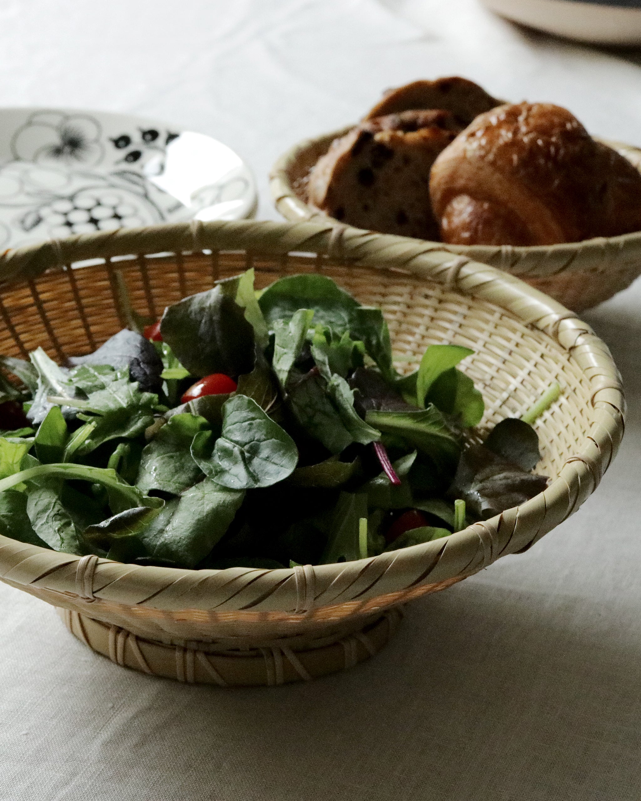 Bowl made out of natural colored woven bamboo with salad in it. There is another bamboo bowl in the background with bread sitting inside. Both bowls are on a beige cloth.