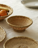 Small bowl made out of natural colored woven bamboo sitting against on a beige cloth. Other woven bowls surround this bowl and one of them has an orange and banana.