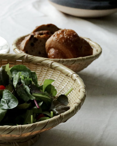 Bowl made out of natural colored woven bamboo with salad in it in the foreground.  There is another bamboo bowl in the background with bread sitting inside. Both bowls are on a beige cloth.