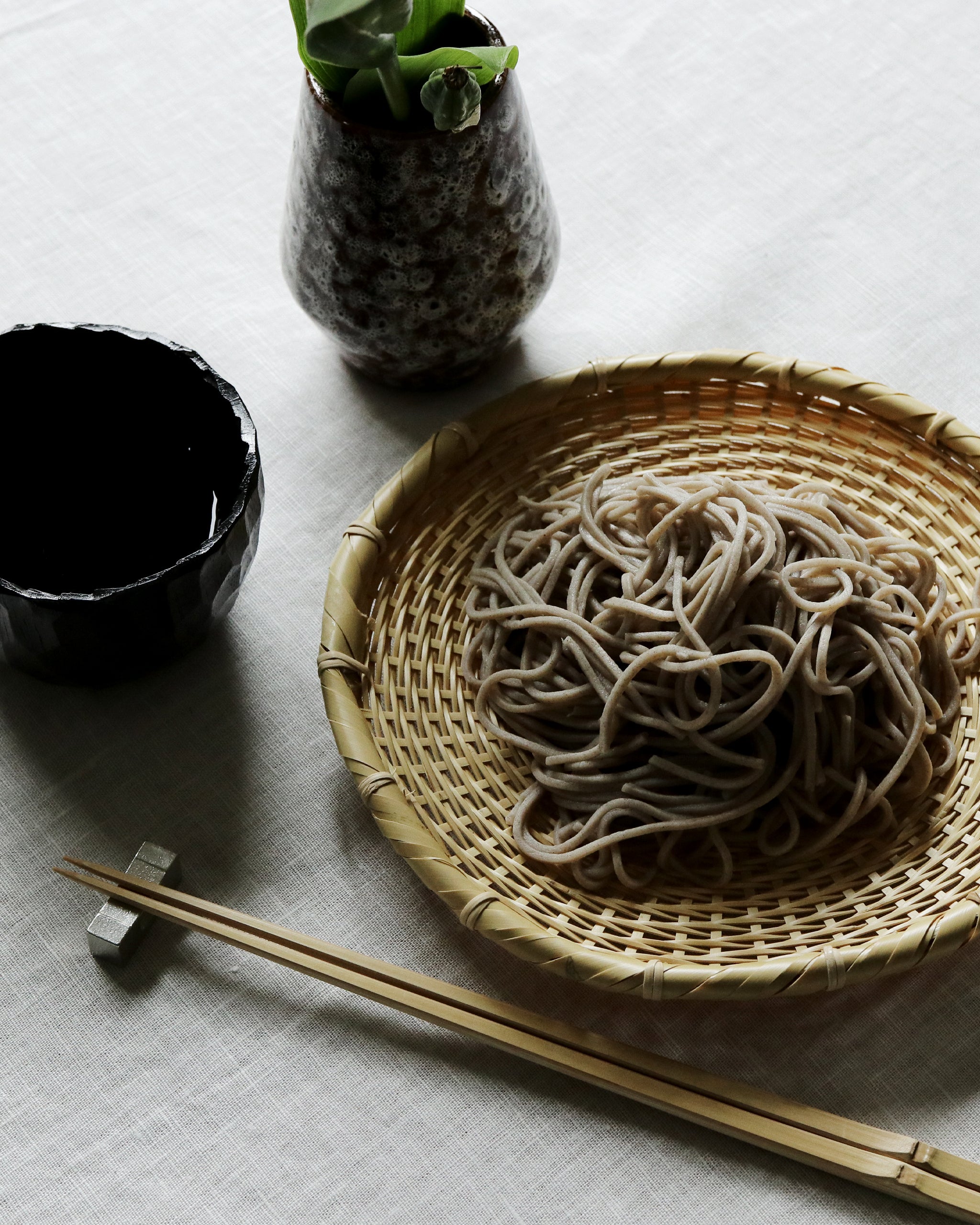 A meal setup. Black cup on the right with another vessel above it. The vessel  is brown and has vegetation in it. On the right of cup and vessel is a woven bamboo tray with soba noodles on it. Below tray is chopsticks on chopstick rest.