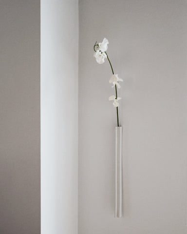 In situation image of ceramic slim white hanging vase with edge with white over glaze by Masanobu Ando against white wall with delicate white flowers.