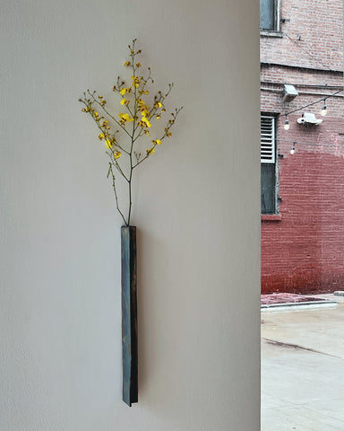 In situation image of ceramic hanging sculpture vase with edge with sterling silver overglaze by Masanobu Ando with delicate yellow flowers mounted on white wall with red brick building in background.