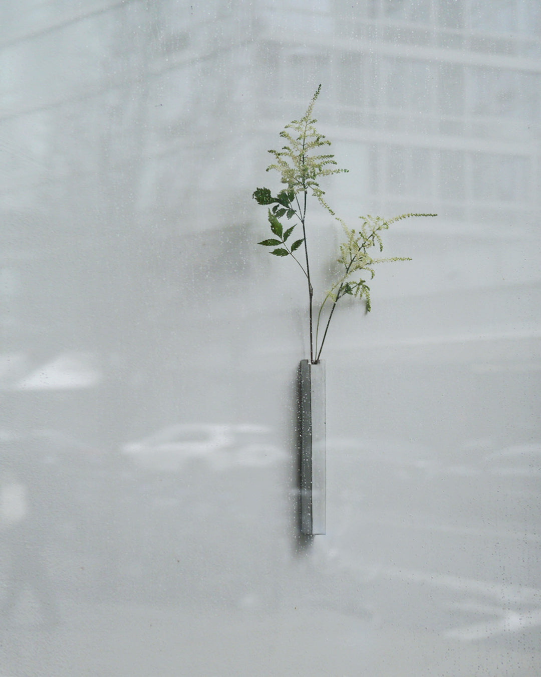 In situation image of short white hanging sculpture vase with white overglaze by Masanobu Ando with delicate white wild flowers behind rainy window.