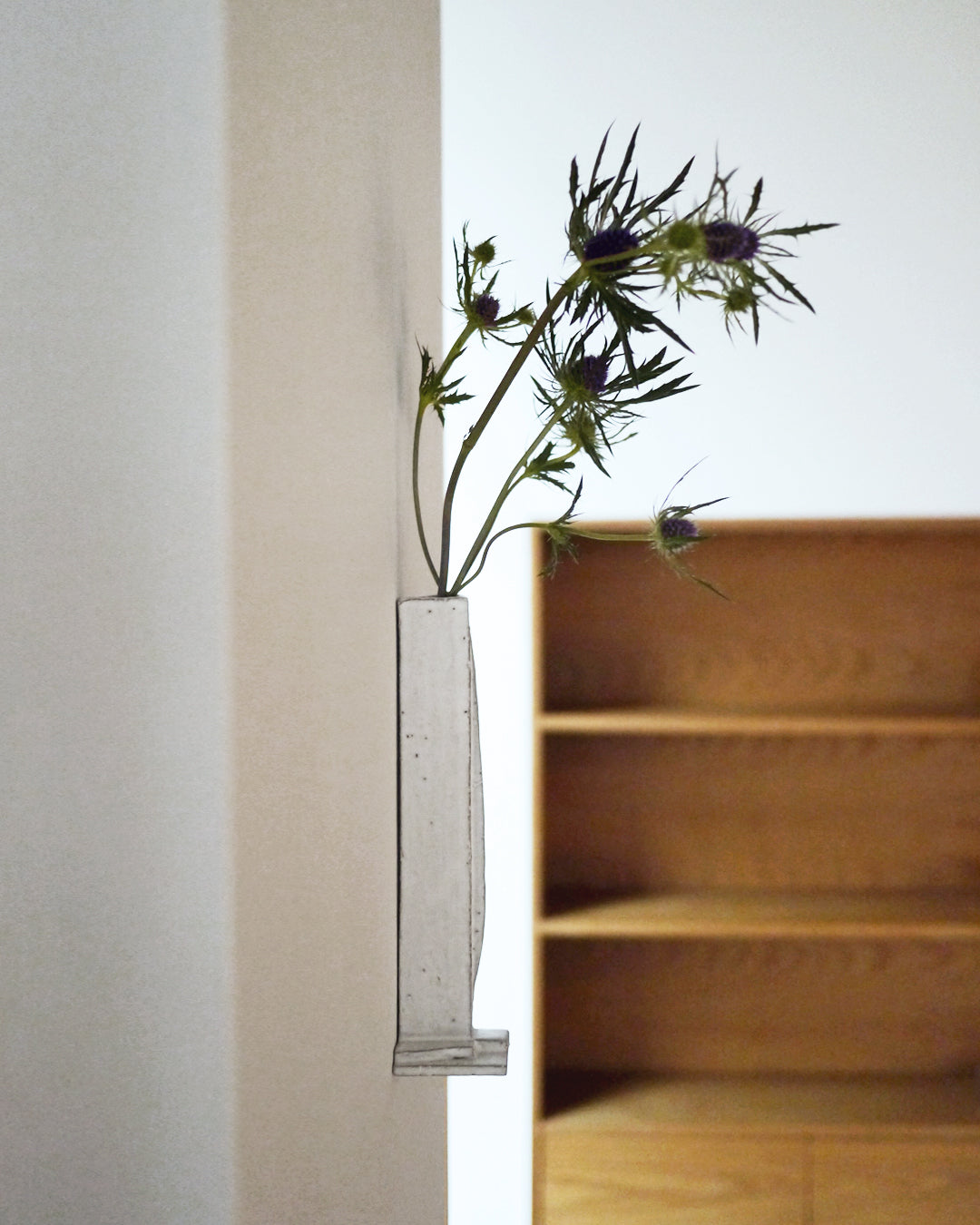 In situation image of ceramic white sculpture vase with foot with white over glaze by Masanobu Ando mounted on white wall with wildflowers and oak bookshelf in background. 