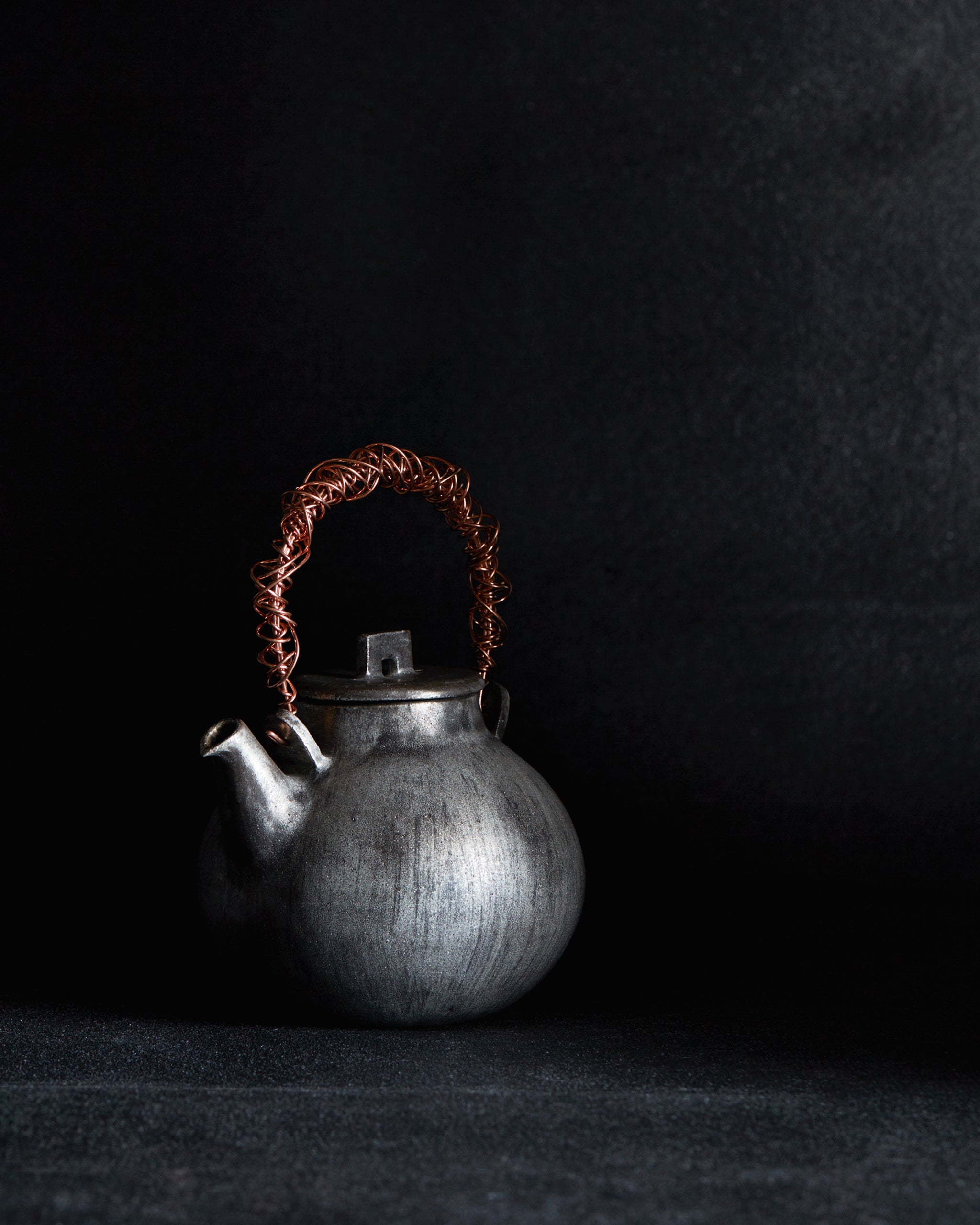 In situation image of small ceramic teapot with sterling silver overglaze and twisted copper wire handle by Masanobu Ando against charcoal-gray background.
