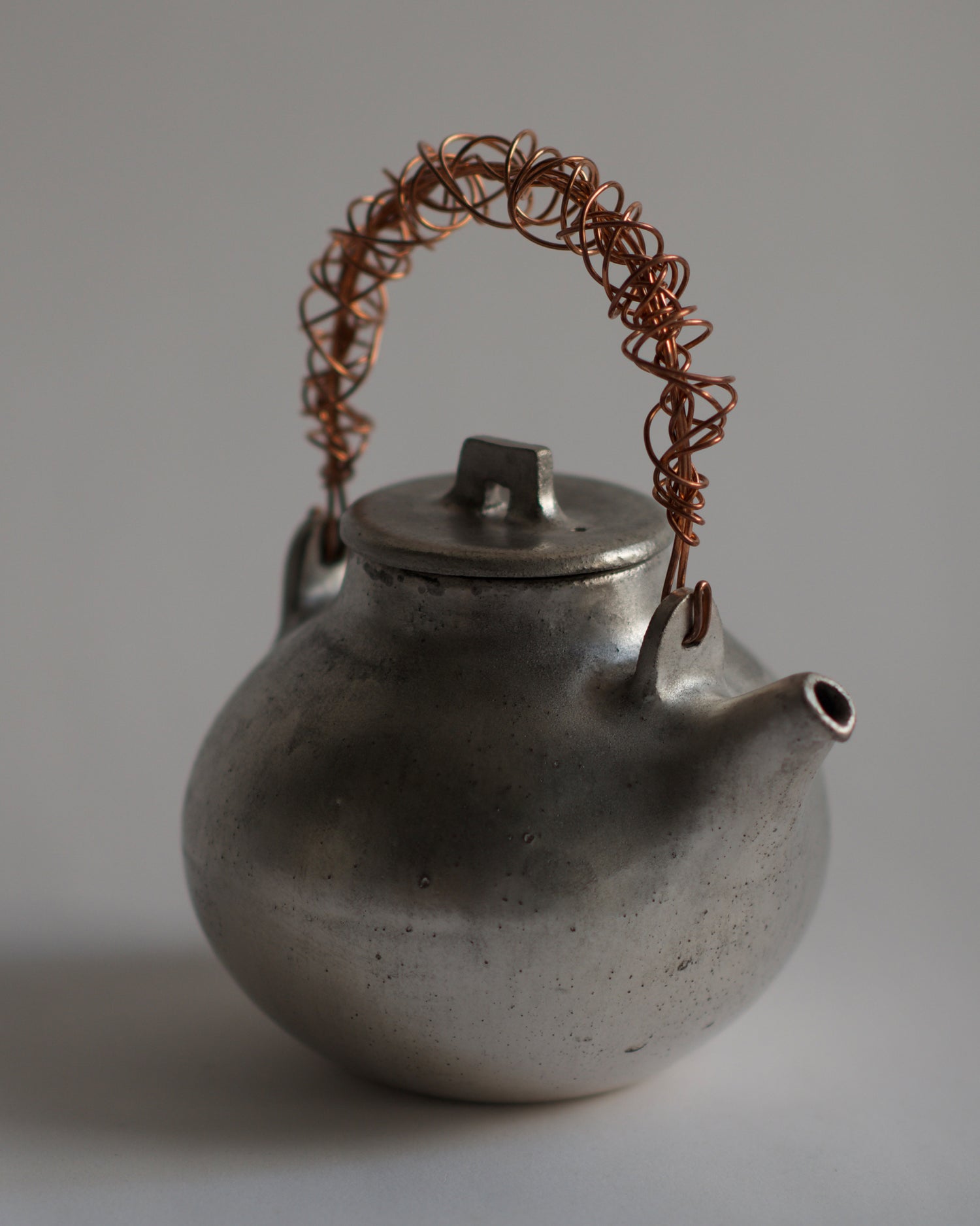 Angled image of small ceramic teapot with sterling silver overglaze and twisted copper wire handle by Masanobu Ando against white-gray background.