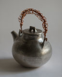 Angled image of back of small ceramic teapot with sterling silver overglaze and twisted copper wire handle by Masanobu Ando against white-gray background.