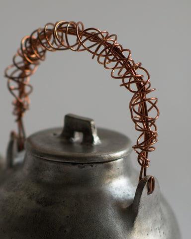 Zoomed-in image of handle of small ceramic teapot with sterling silver overglaze and twisted copper wire handle by Masanobu Ando against white-gray background.