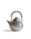 Silhoutted image of small ceramic teapot with sterling silver overglaze and twisted copper wire handle by Masanobu Ando against white background.