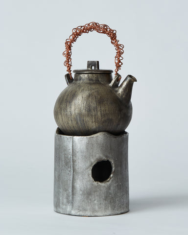 In situation image of ceramic teapot with silver overglaze and twisted copper handle on top of ceramic teapot warmer with sterling silver overglaze by Masanobu Ando against white-gray background.