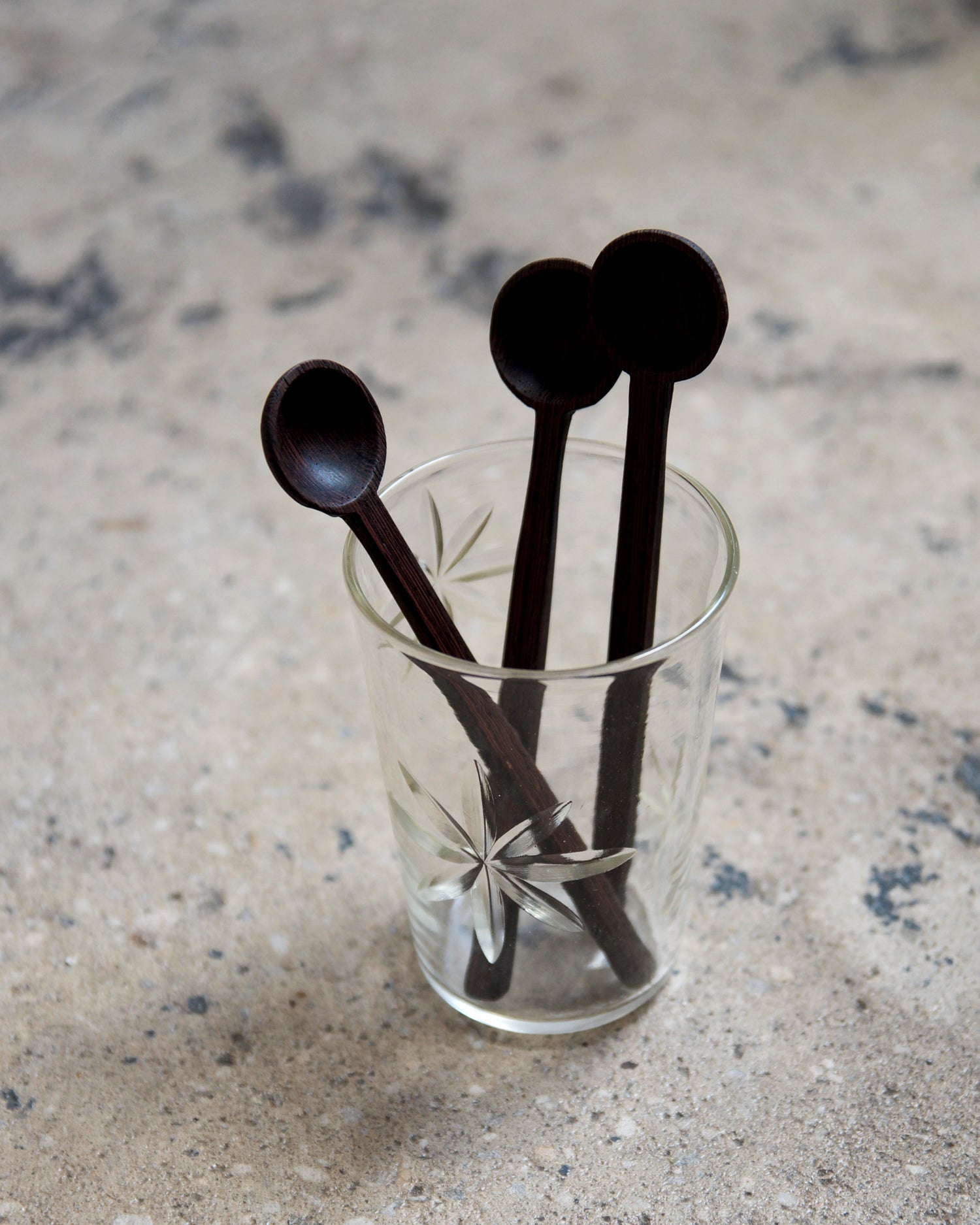 In situation image of three hand carved tagaya wood spoons by Nalata Nalata in a carved glass cup on a cement surface. 