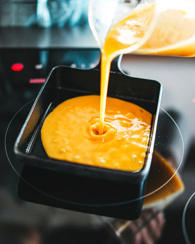 Pitcher pouring yellow egg mixture into a cast iron pan on a stovetop.