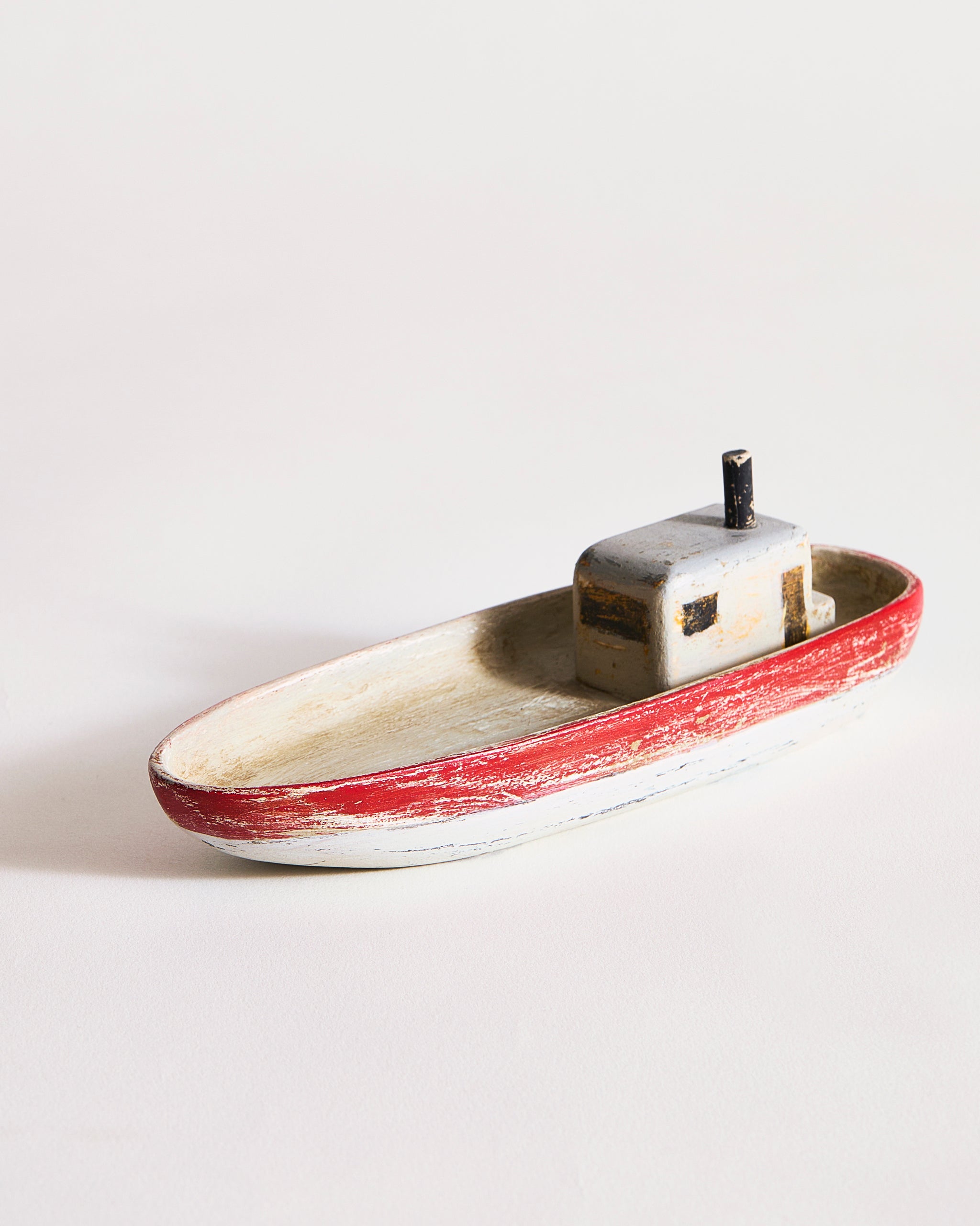 Small wooden boat with red,gray, and black paint by Ryuji Mitani on a light gray background