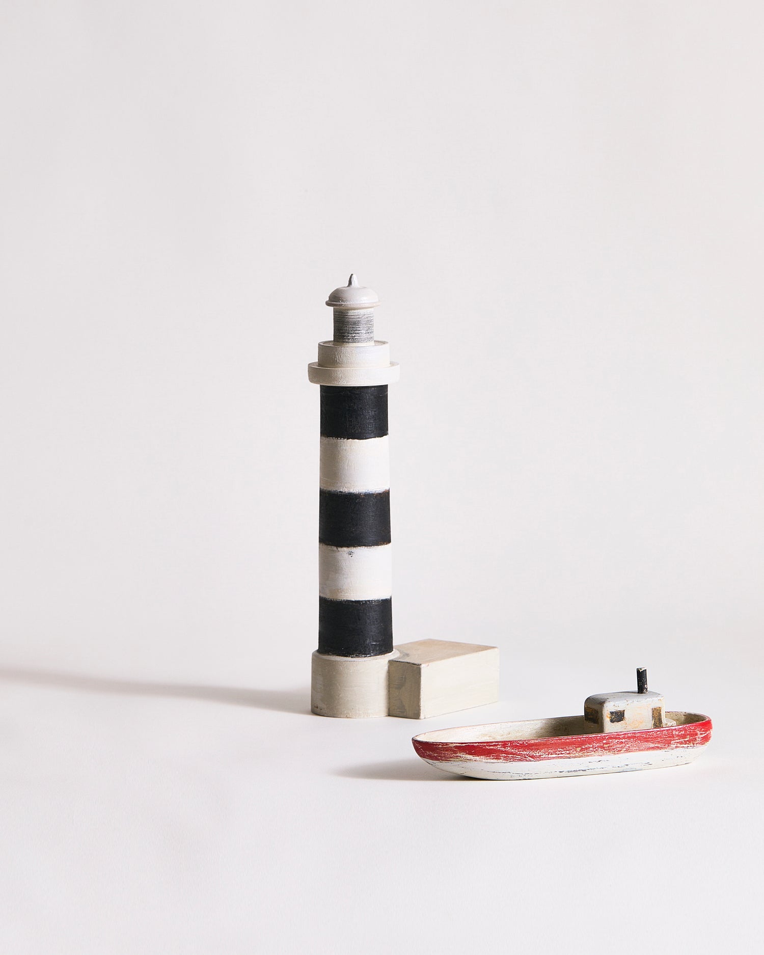 Wooden lighthouse and small wooden boat with red, gray, and black paint by Ryuji Mitani on a light gray background