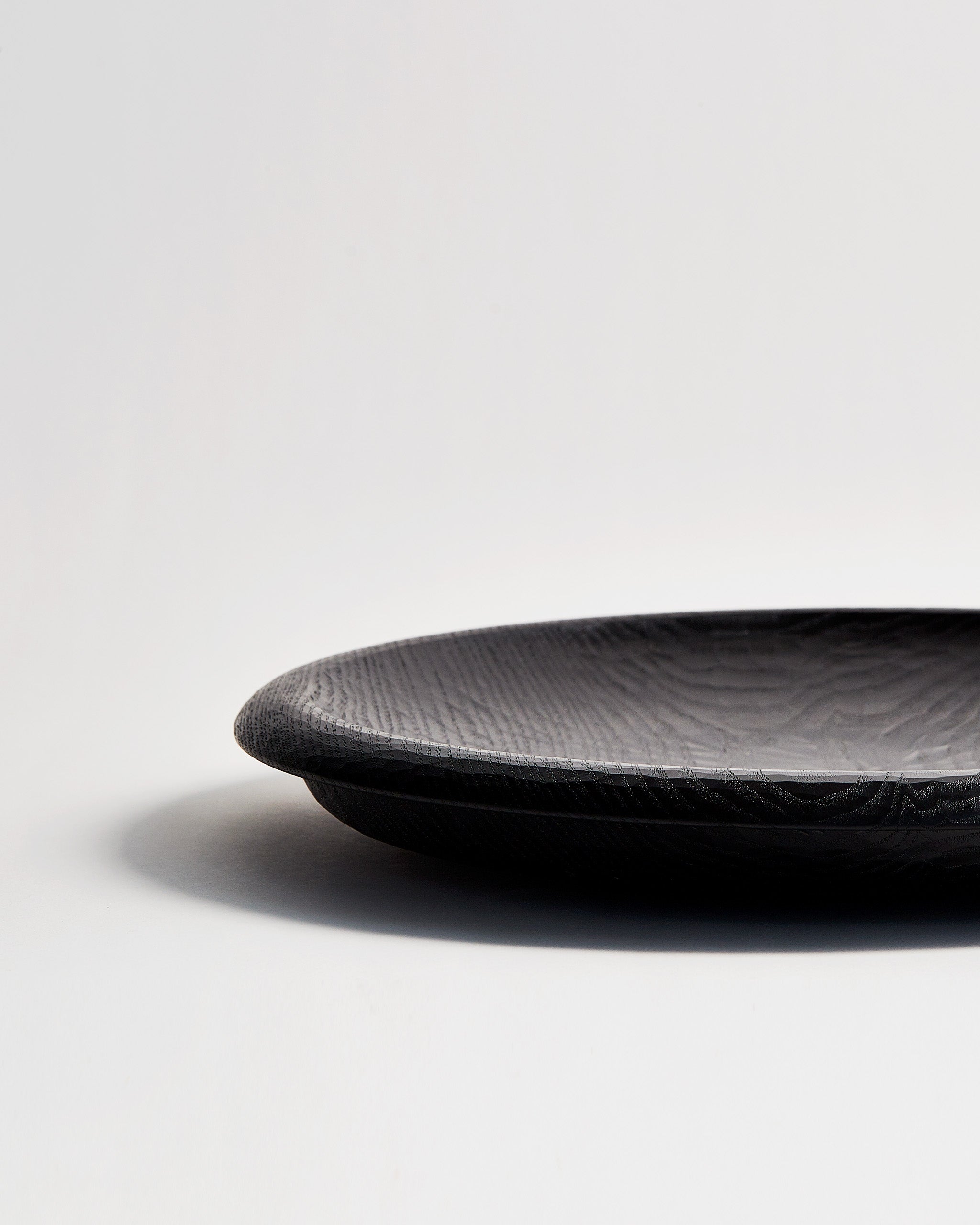 Cropped view of Noir Round Curved Rim Platter by Ryuji Mitani against white-gray background. 