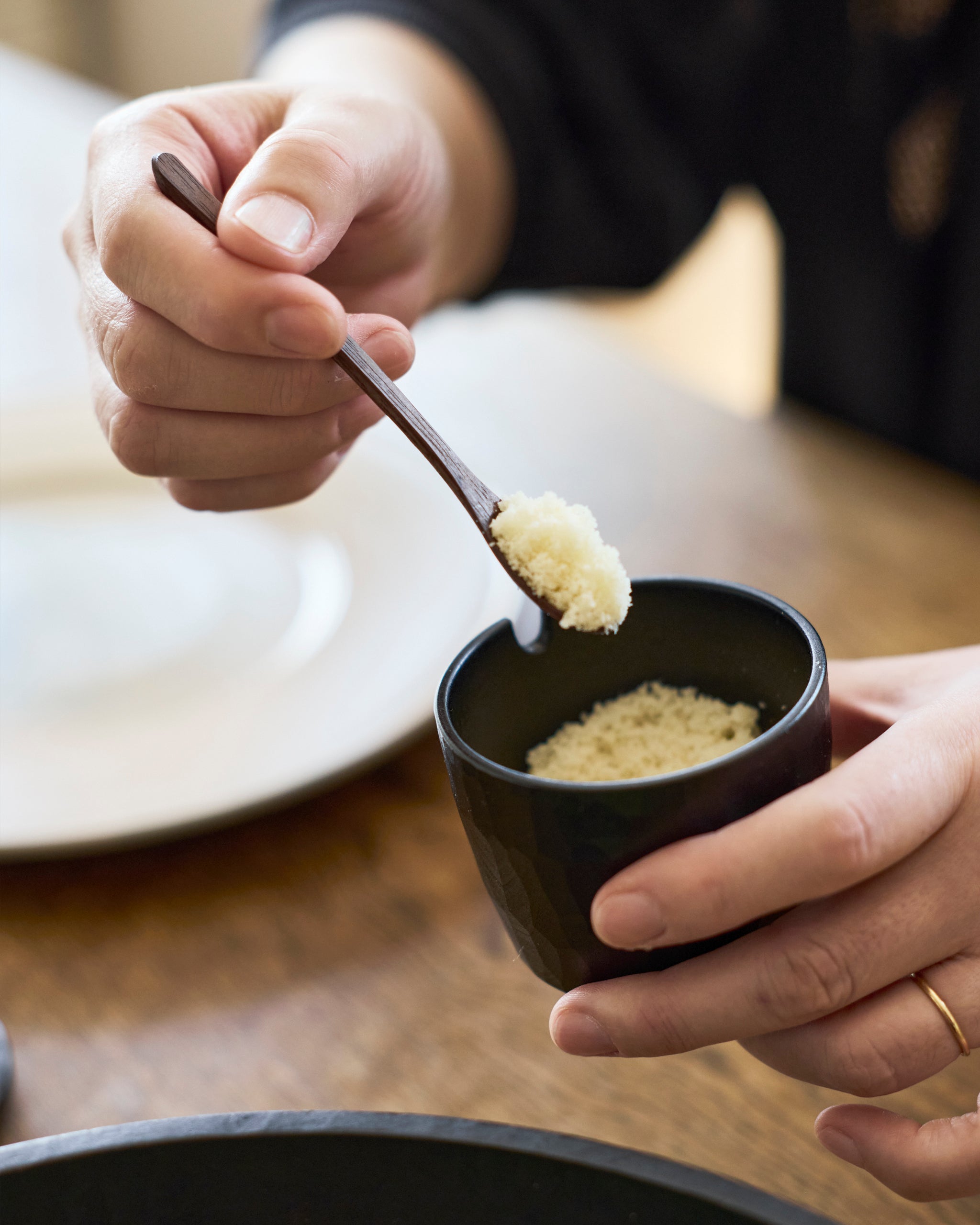 Person holding a small black spice container with a wooden spoon made by Ryuji Mitani scooping something out.