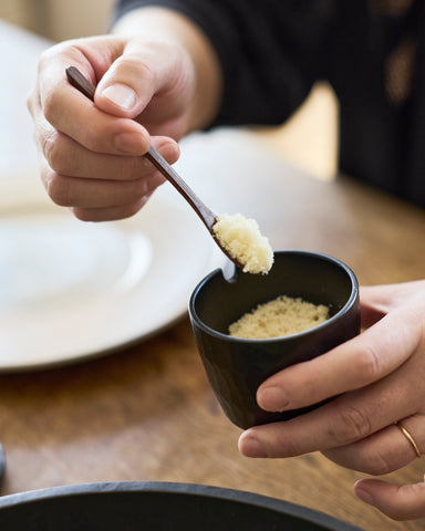 Person holding a small black spice container with a wooden spoon made by Ryuji Mitani scooping something out.
