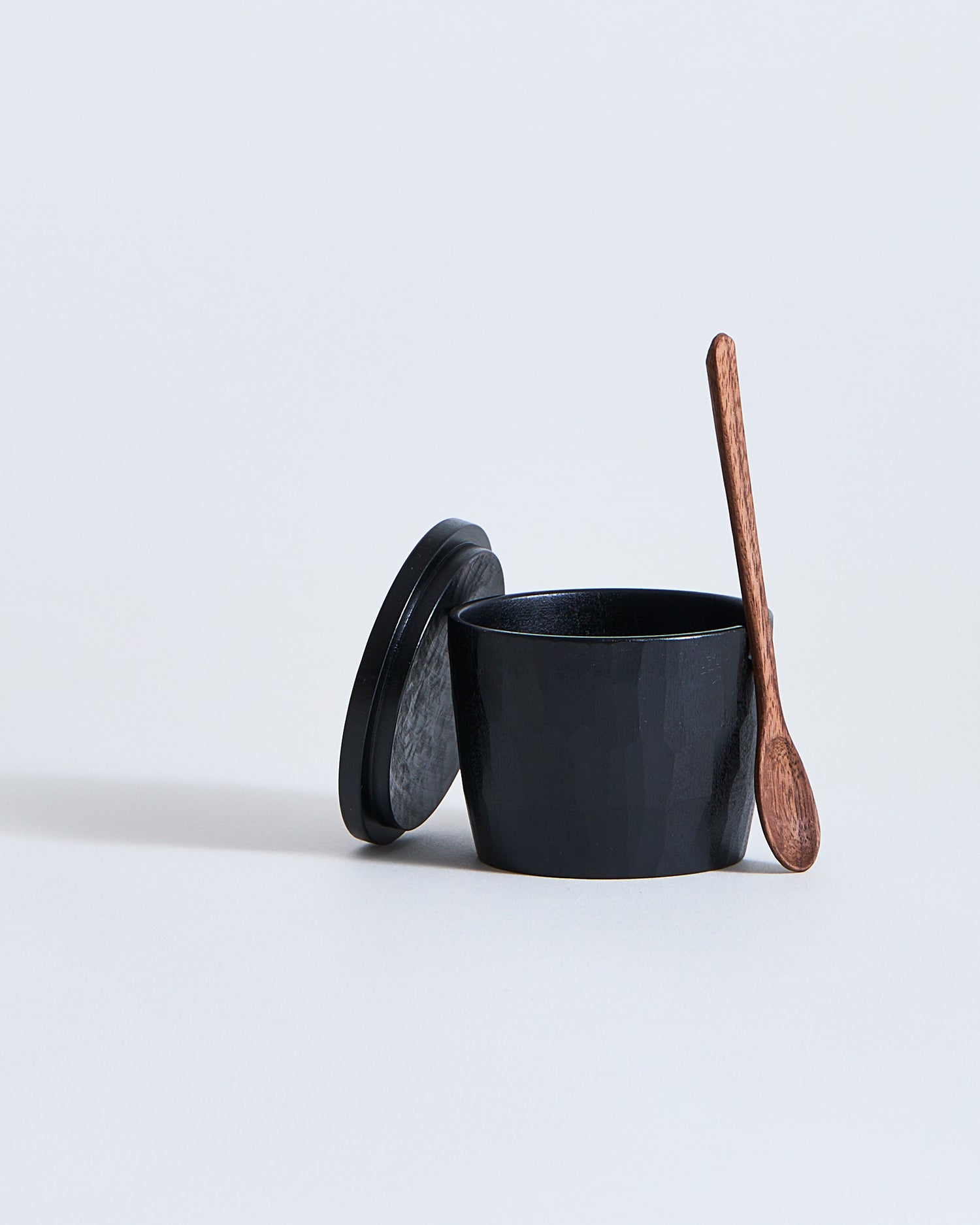 Black wooden spice container with wooden lid and spoon by Ryuji Mitani on a light gray background