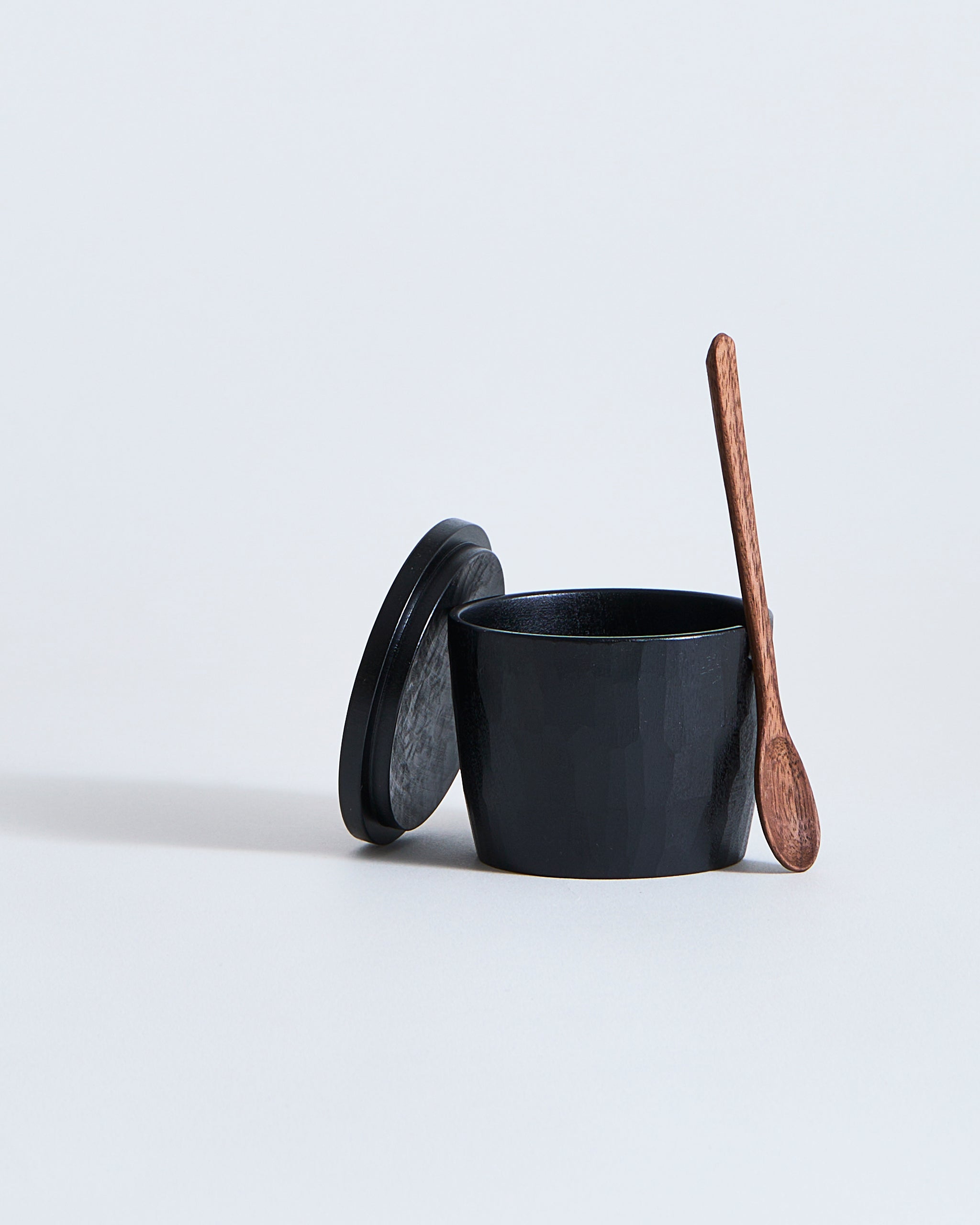 Black wooden spice container with wooden lid and spoon by Ryuji Mitani on a light gray background