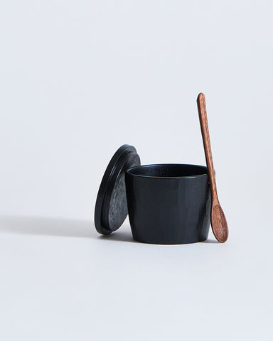 Black wooden spice container with wooden lid and spoon by Ryuji Mitani on a light gray background