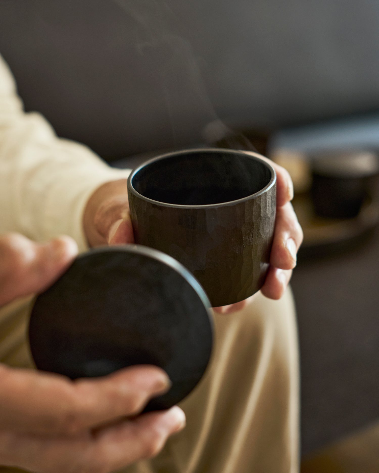 Person holding a dark wooden lid and cup with steam rising, blurred background