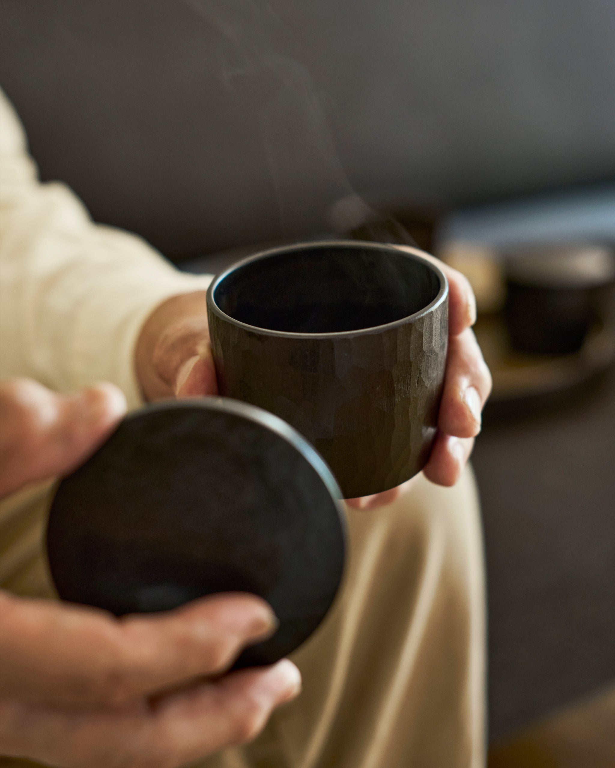 Person holding a dark wooden lid and cup with steam rising, blurred background